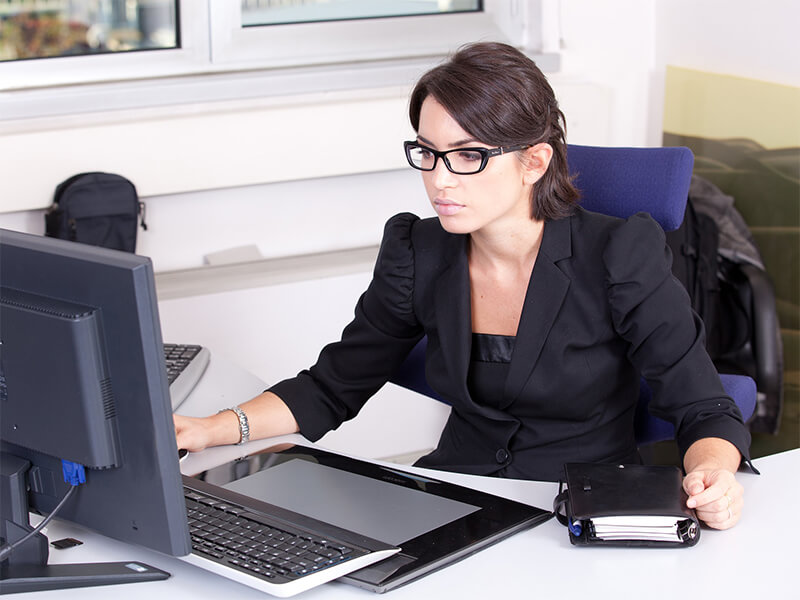 Millennial woman with brown hair and glasses wearing a black blouse looking at her desktop computer monitor